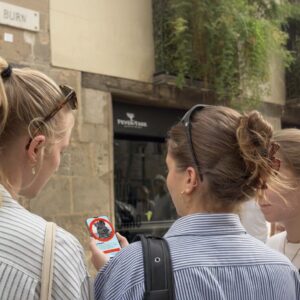 Three young women stand on a street in Barcelona's El Born district, looking at a city game on a smartphone.