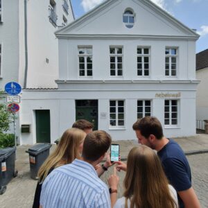 A group of friends playing a city exploration game on a smartphone in front of a white building in Bielefeld.