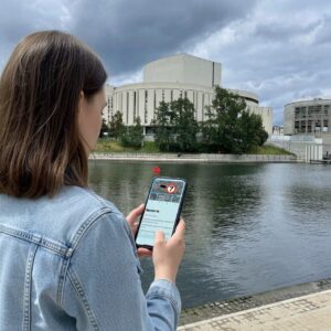 A woman plays a city game on her phone by the river in Bydgoszcz, with the Opera Nova building behind her.