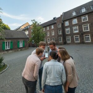 A group of six young adults look at a smartphone together in the cobblestone courtyard of the Stadtmuseum in Dusseldorf.