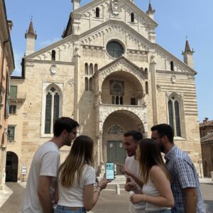 A group of five young friends stand together looking at a smartphone in front of the Duomo di Verona.