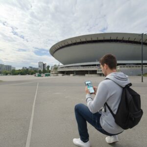 A young man in a grey hoodie crouches with a phone in front of the Spodek arena in Katowice.