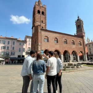 A group of five young people look at a smartphone in front of the historic Arengario in Monza.