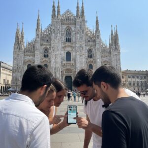 A group of five young people gathered around a smartphone in front of the Milan Cathedral.