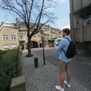 A young man with a backpack stands on a cobblestone street in Roermond using a tablet.