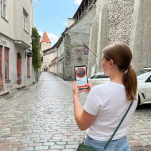 A woman stands on a cobblestone street in Tallinn's Old Town holding a phone with a city game.
