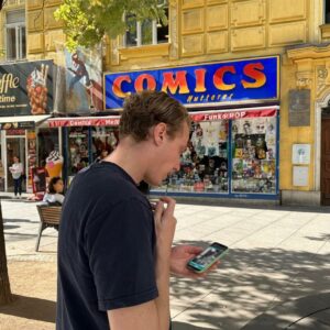 A young man playing a city game on his phone in front of the Comics Hutterer shop in Vienna.