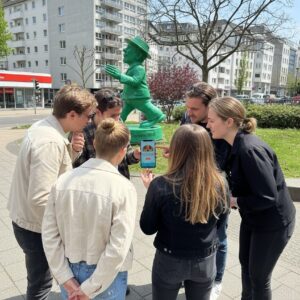 A group of young people gather around a smartphone next to the green Mainzelmännchen statue in Wiesbaden.