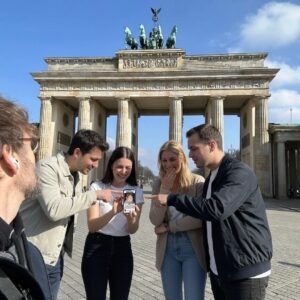 A group of friends smiles and points at a smartphone while standing before the Brandenburg Gate in Berlin.
