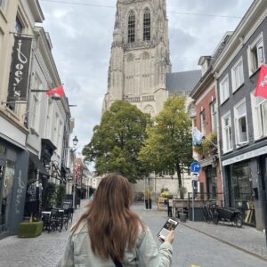 A woman holding a phone walks down a historic street in Breda towards the Grote Kerk church tower.