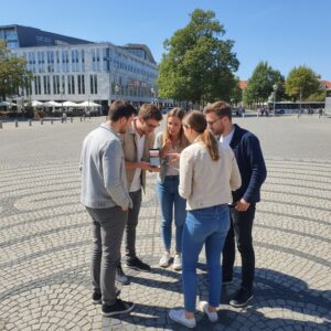 A group of five friends stands together on the Universitätsplatz in Magdeburg, looking at a smartphone.