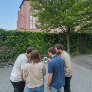 A group of four people play a city game on a smartphone near the historic Stadtkirche in Darmstadt.