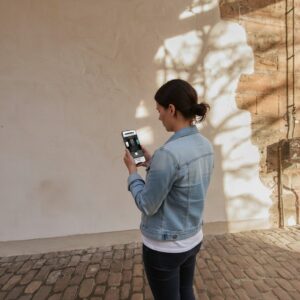 A woman in a denim jacket stands on a cobblestone street in Freiburg im Breisgau, playing a game on her smartphone.