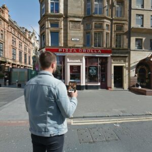 A man in a denim jacket stands on a street corner in Glasgow using his phone.