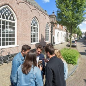A group of five friends stand on a historic cobblestone street in Harderwijk, looking at a smartphone.