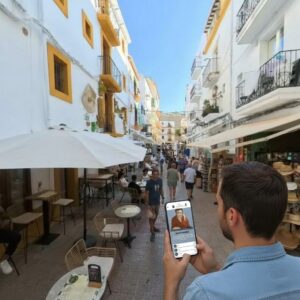 A man holds a smartphone displaying a game while walking down a busy, narrow street in Ibiza.