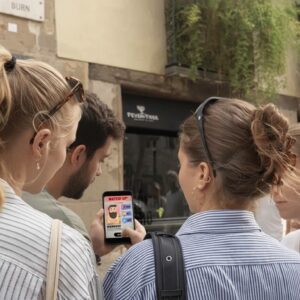 A group of friends stand together on a city street in Kefalonia looking at a game on a smartphone.