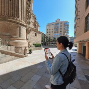 A woman plays a city exploration game on her phone in a sunny square next to the Málaga Cathedral.