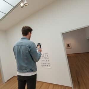 A man wearing a denim jacket plays a city game on his phone inside a modern art museum in Maastricht.