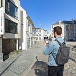 A man with a backpack stands on a cobblestone street in Bergen, playing a city game on a tablet.