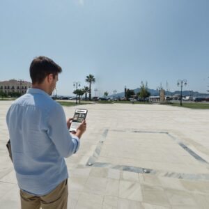 A man stands in the sunny Solomos Square in Zakynthos, looking at a tablet with the harbor behind.