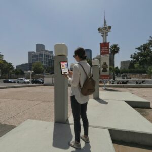 A woman stands in a plaza holding a tablet in front of the Tel Aviv Museum of Art.