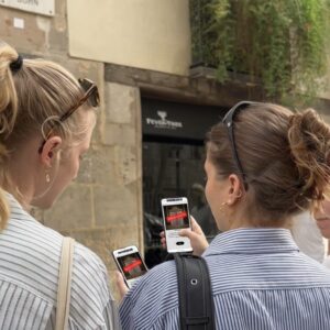 Three young women stand together on a street in Samos, looking at smartphones while playing a city game.