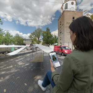 A woman plays a city game on her phone in the market square of Emmen with the Grote Kerk tower behind her.