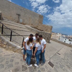 A group of five young people in white shirts and jeans gather around a smartphone in Ibiza.