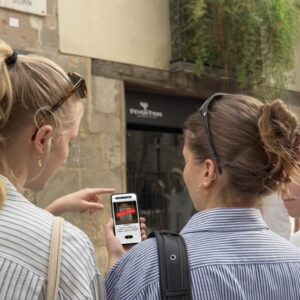 Three young women stand on a street in Faro, looking at a smartphone displaying a Sherlock game.