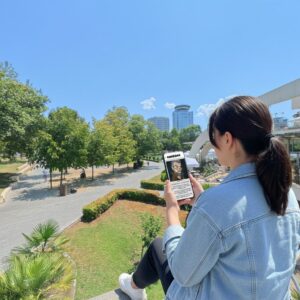 A woman plays the Sherlock city game on her phone in a sunny park in Tirana.