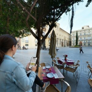 A woman sits at an outdoor cafe table playing a city game on her phone in Valencia.