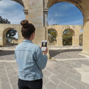 A woman plays a game on her phone under the historic stone arches of the Upper Barrakka Gardens in Valletta.