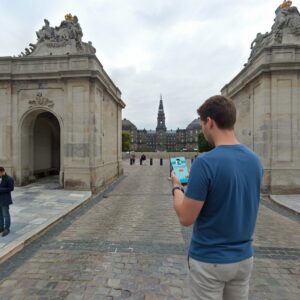 A man looks at his phone while standing between two gateways facing Christiansborg Palace in Copenhagen.