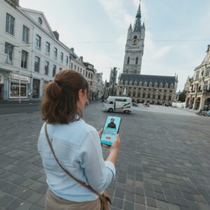 A woman uses a smartphone to play a city game in a square before the Ghent Belfry.