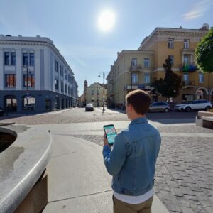 A young man in a denim jacket stands in Rūdninkų Square in Vilnius, looking at his smartphone.