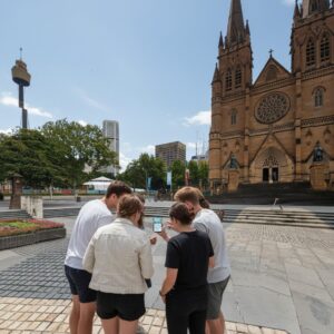 A group of five people gather around a smartphone in a plaza with St Mary's Cathedral in Sydney.