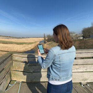 A woman in a denim jacket holds a smartphone while looking out over the polder landscape of Zaanse Schans.